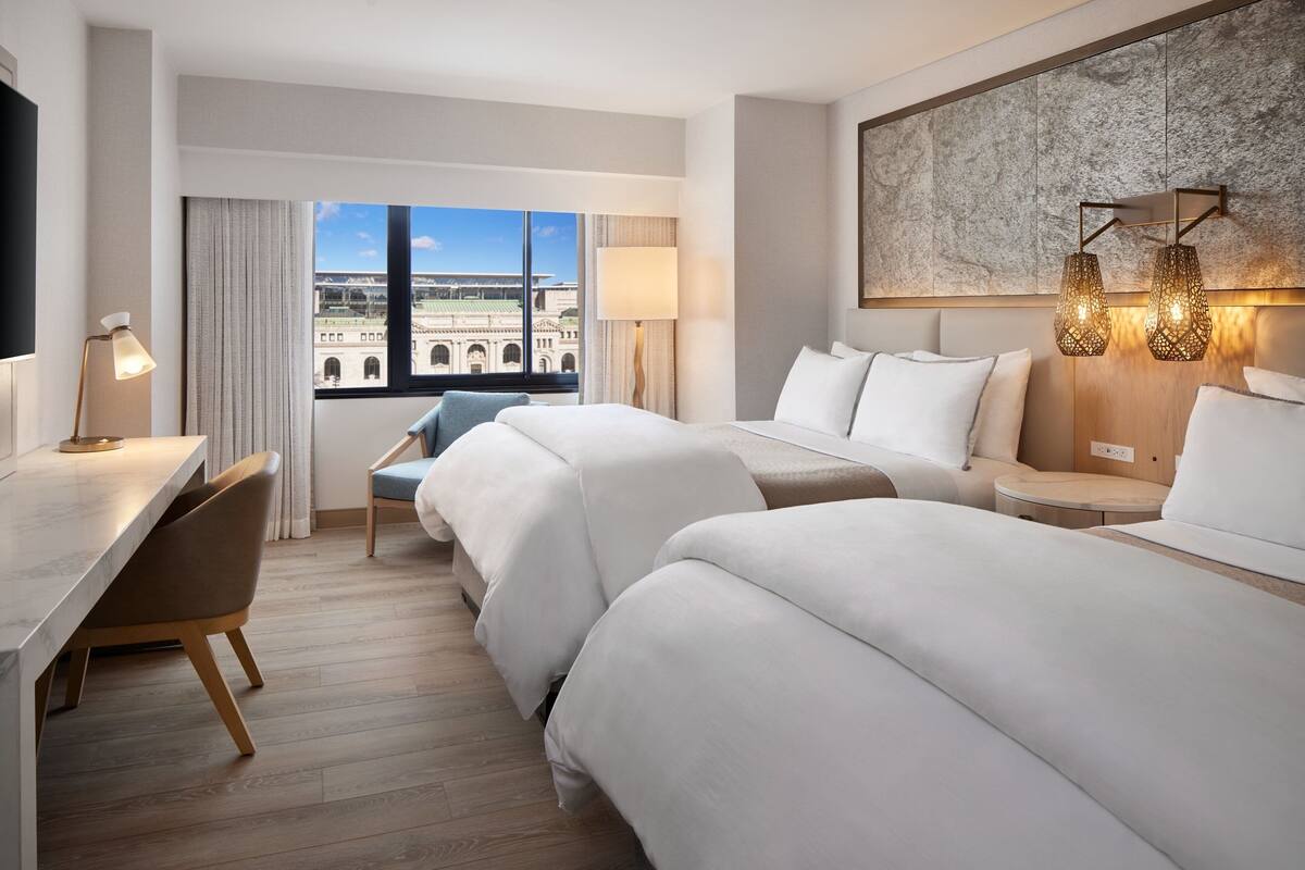A hotel room with two beds, a desk, a chair, and a large window offering a view of a building.