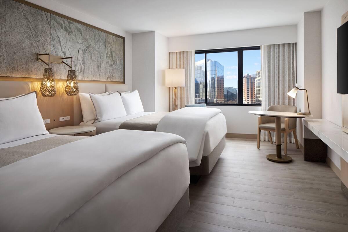 A hotel room with two beds, a desk, a chair, a lamp, and a view of the city.