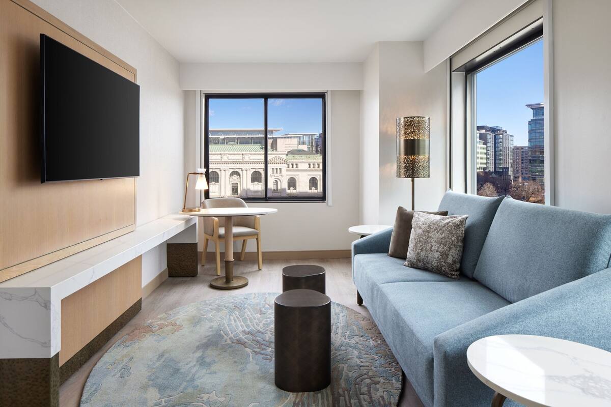 A modern hotel room with a large window offering a city view, a flat-screen TV mounted on the wall, a blue sofa, a small round table, and a decorative floor lamp.