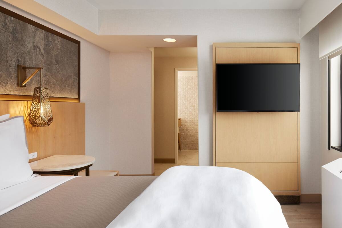 A modern hotel room with a bed, a flat-screen TV mounted on a wooden panel, a small table with a chair, and a decorative wall piece.