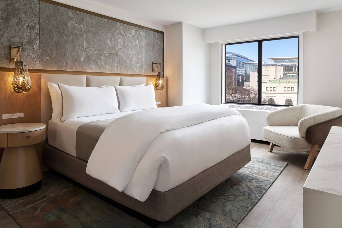 A modern hotel room with a large bed, a bedside table, a chair, and a view of the city through a large window.
