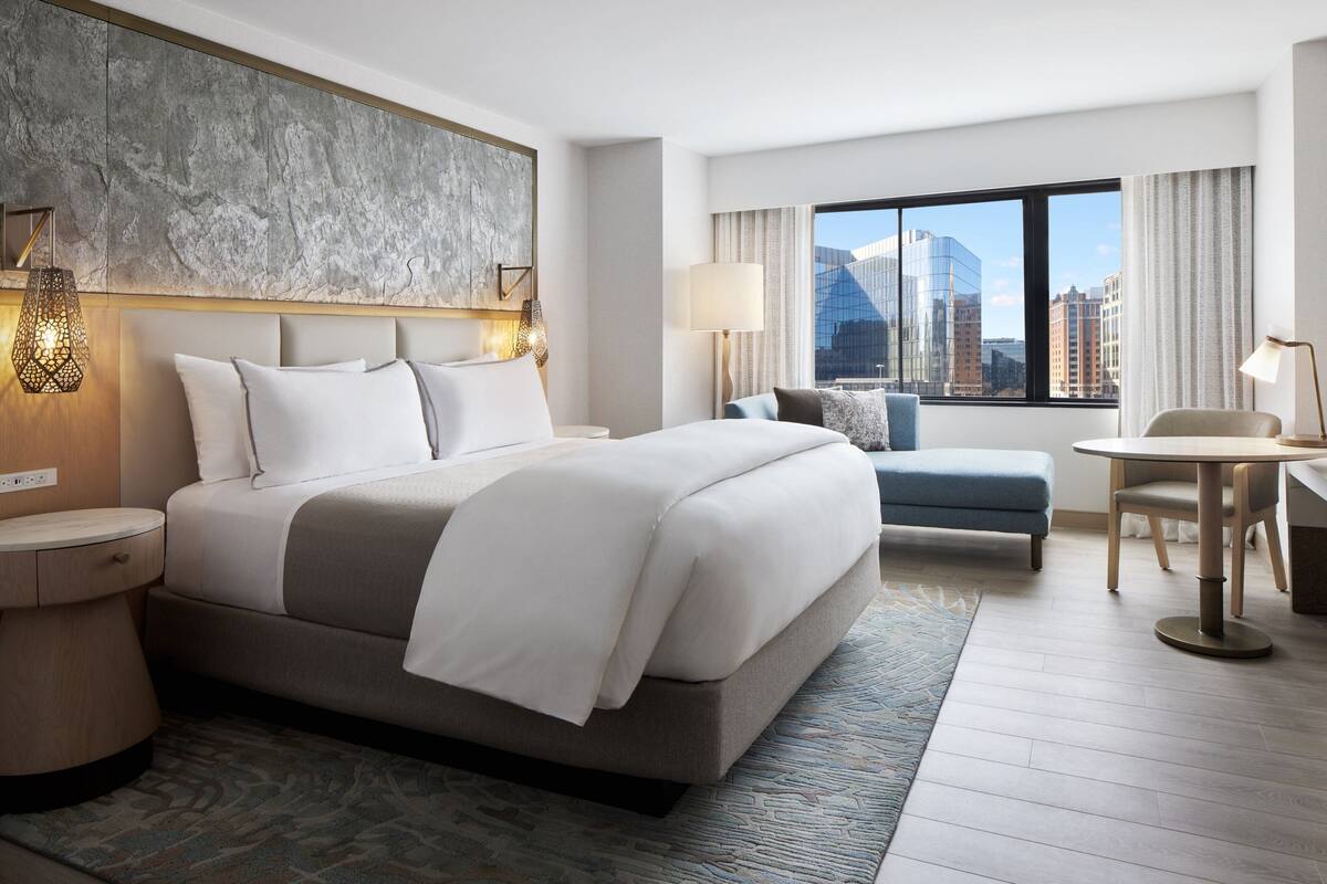A modern hotel room with a large bed, a seating area, a dining table, and a city view through the window.