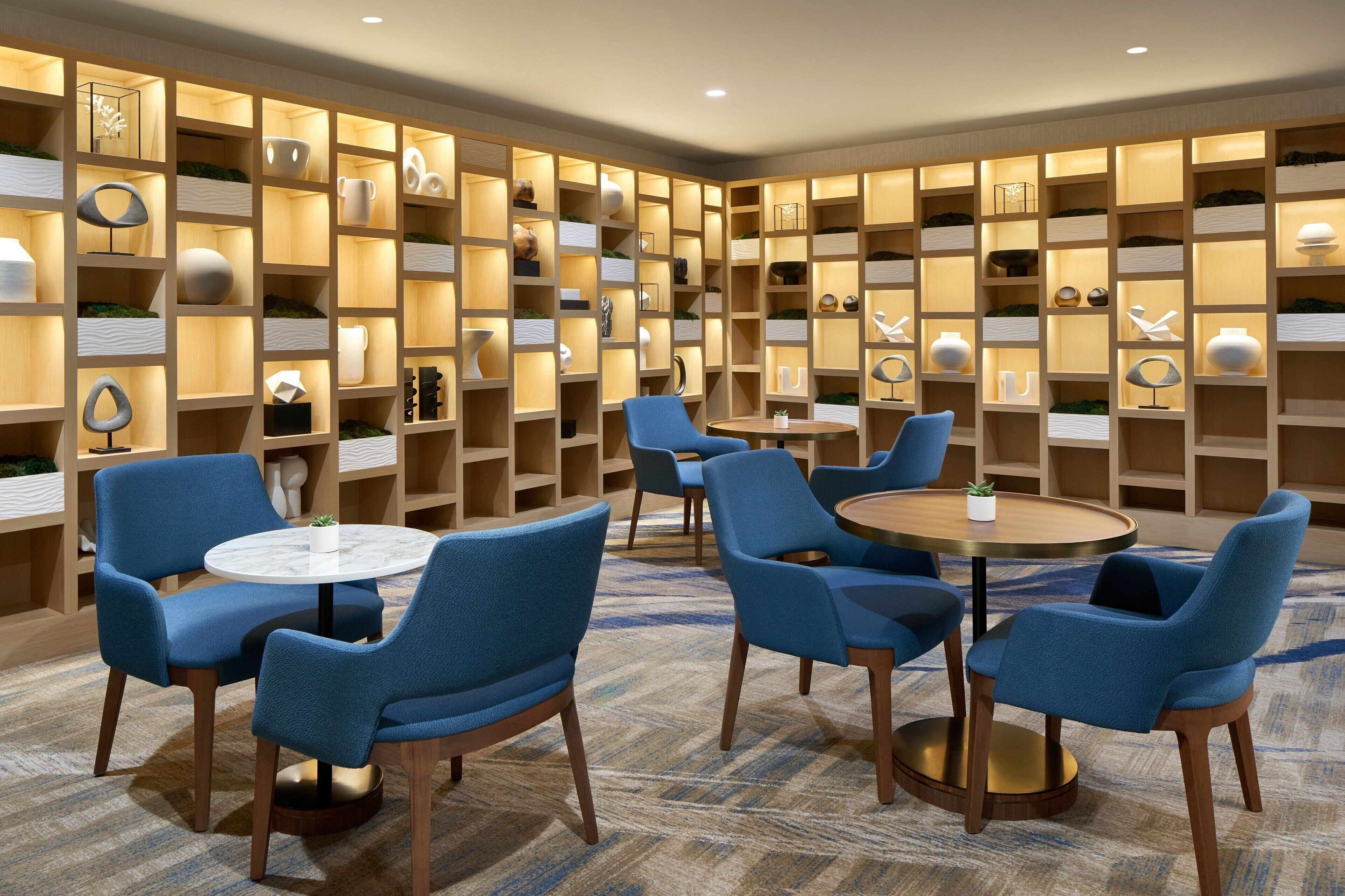 Um interior moderno com cadeiras estofadas em azul e mesas redondas, cercado por estantes de madeira com diversos objetos decorativos.