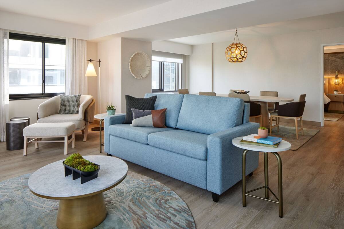 A modern living room with a blue sofa, a round coffee table with a plant, a dining area with a table and chairs, and a large window with curtains.