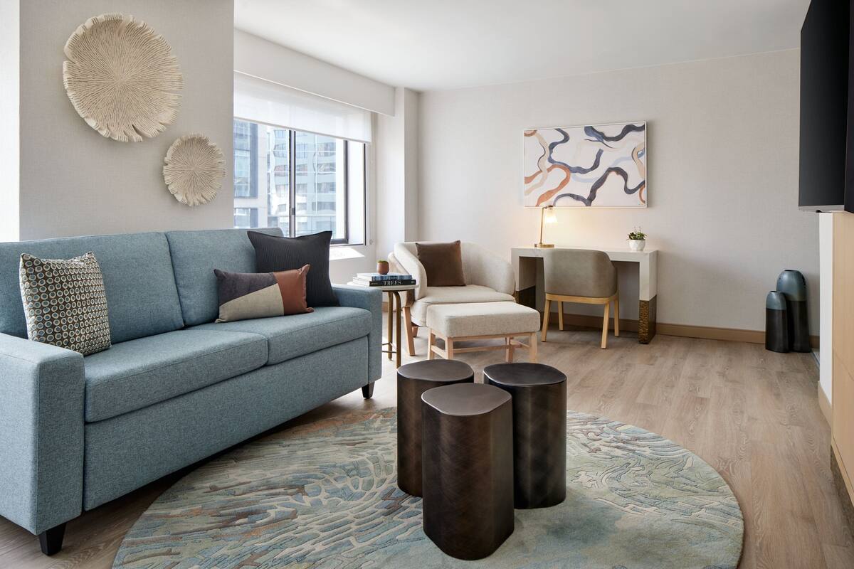A modern living room with a blue sofa, a round rug, and a coffee table. There is a painting on the wall and a window with a view of buildings.