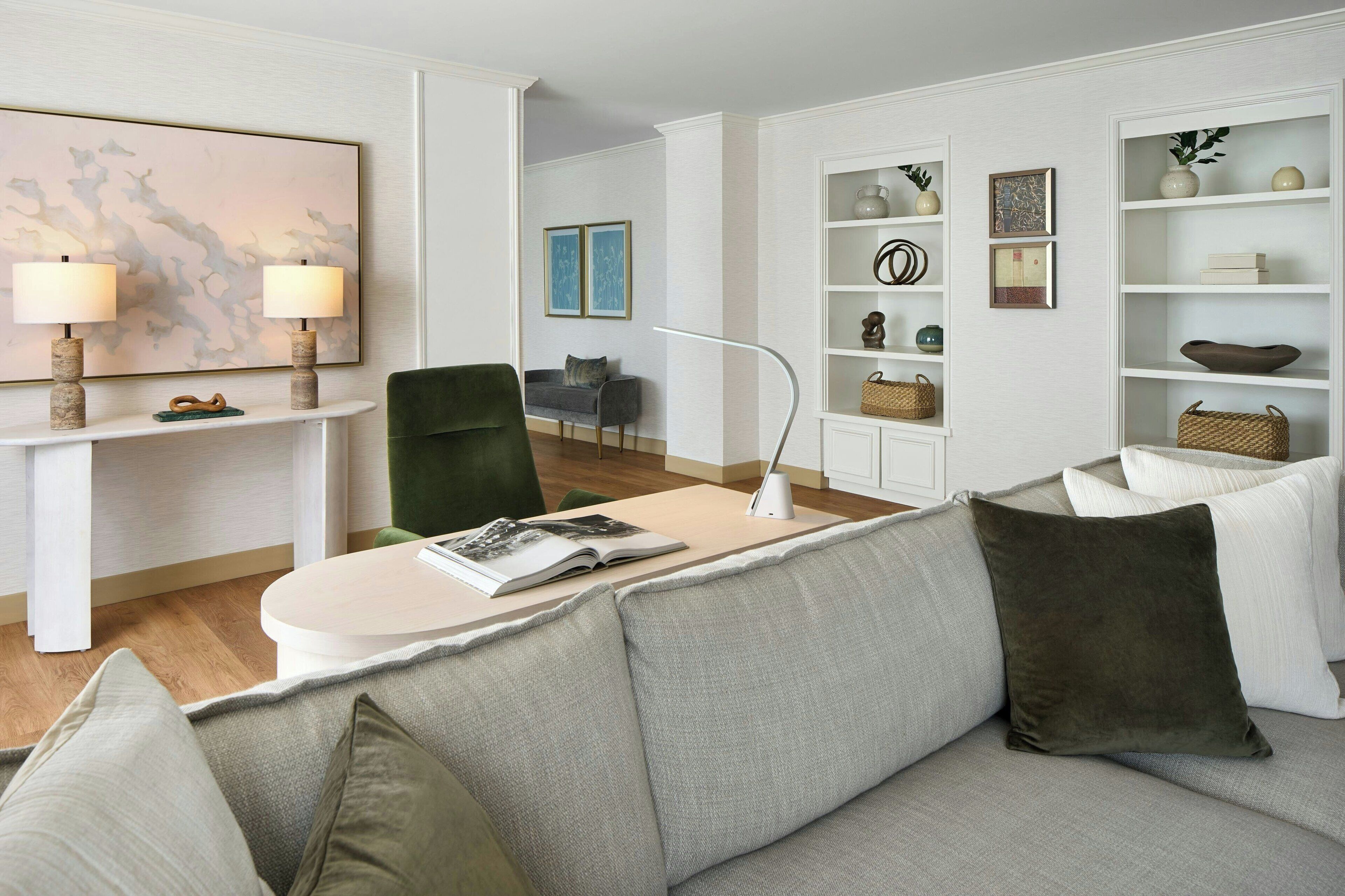 A modern living room with a grey sofa, a coffee table with a book, a side table with lamps, and a bookshelf with decorative items.