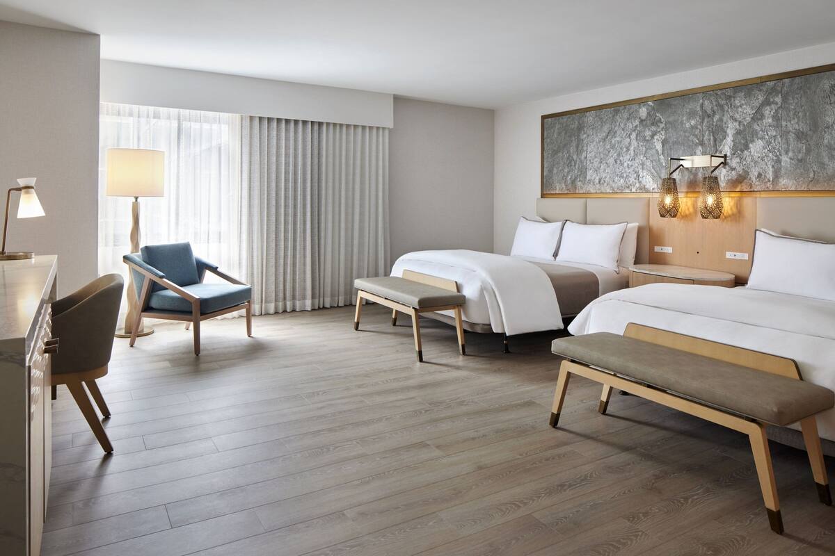A hotel room with a large bed, two bedside tables, a bench, a desk with a lamp, a chair, and a large window with curtains.