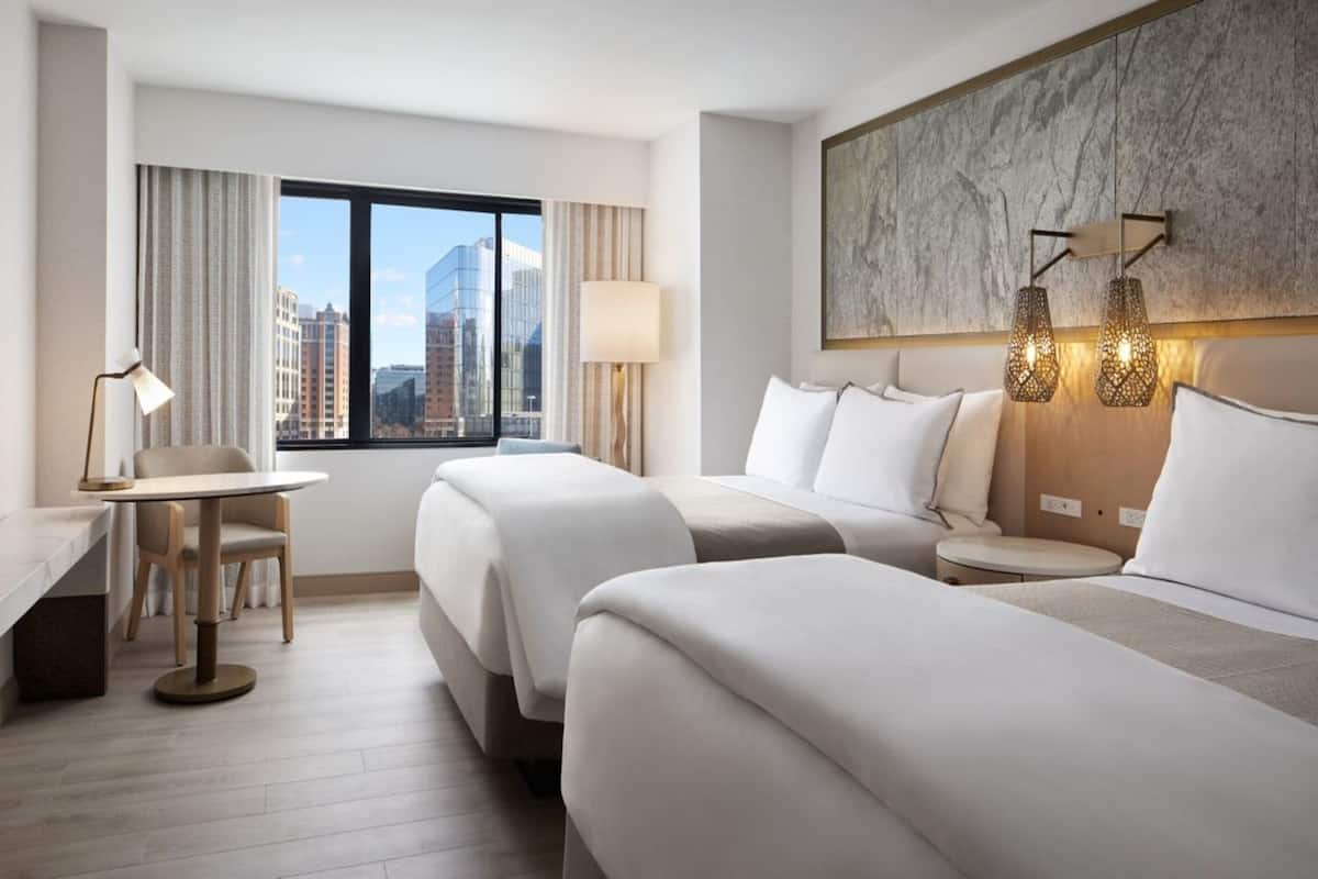 A hotel room with two beds, a desk, a chair, a lamp, and a view of the cityscape.
