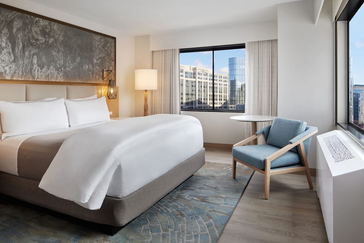 A modern hotel room with a large bed, a seating area with a chair, a small table, and a view of the city through a large window.