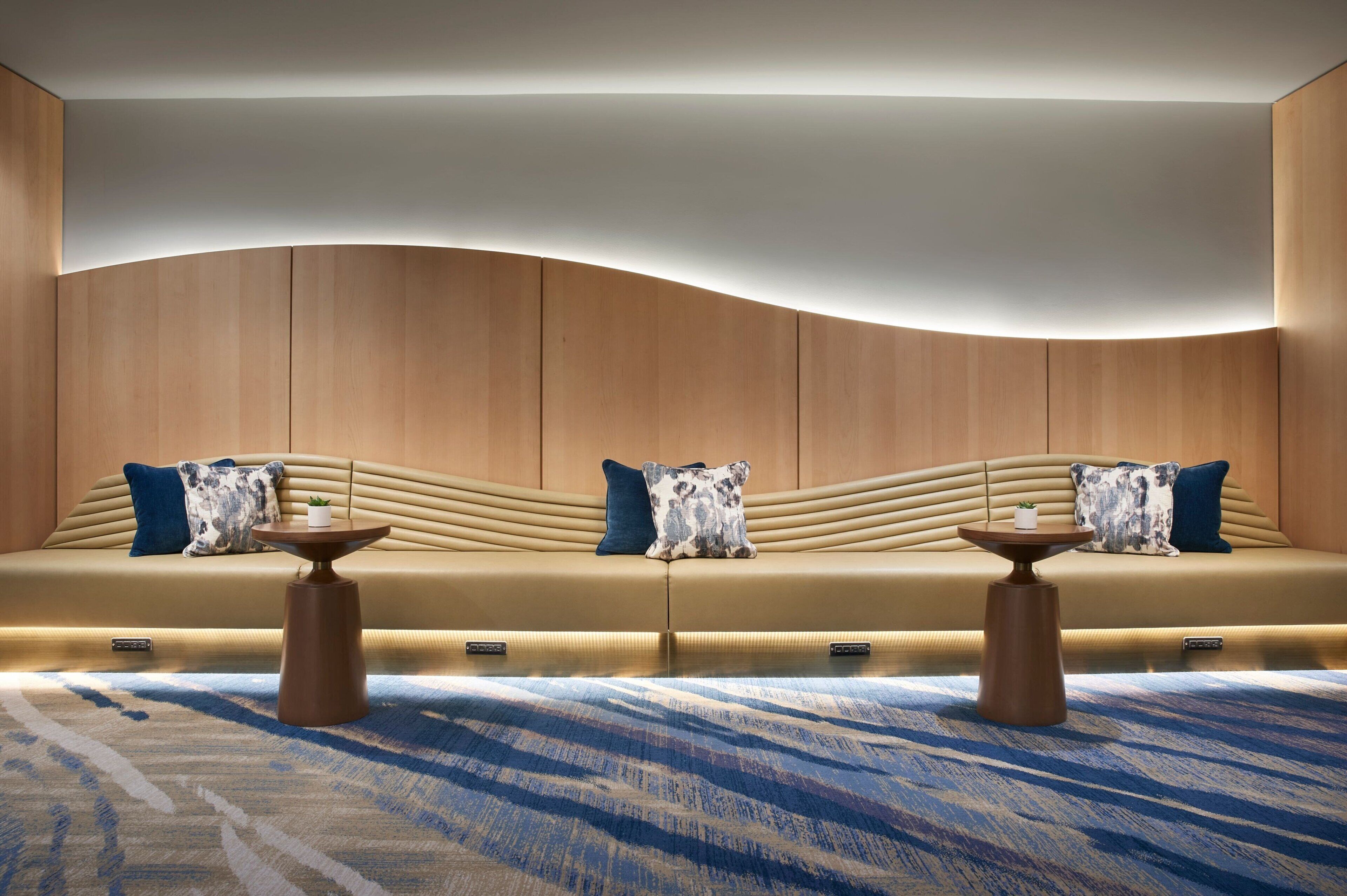 Une pièce moderne et bien éclairée, avec un mur en bois aux lignes courbes, un canapé beige agrémenté de coussins à motifs et deux petites tables rondes en bois.