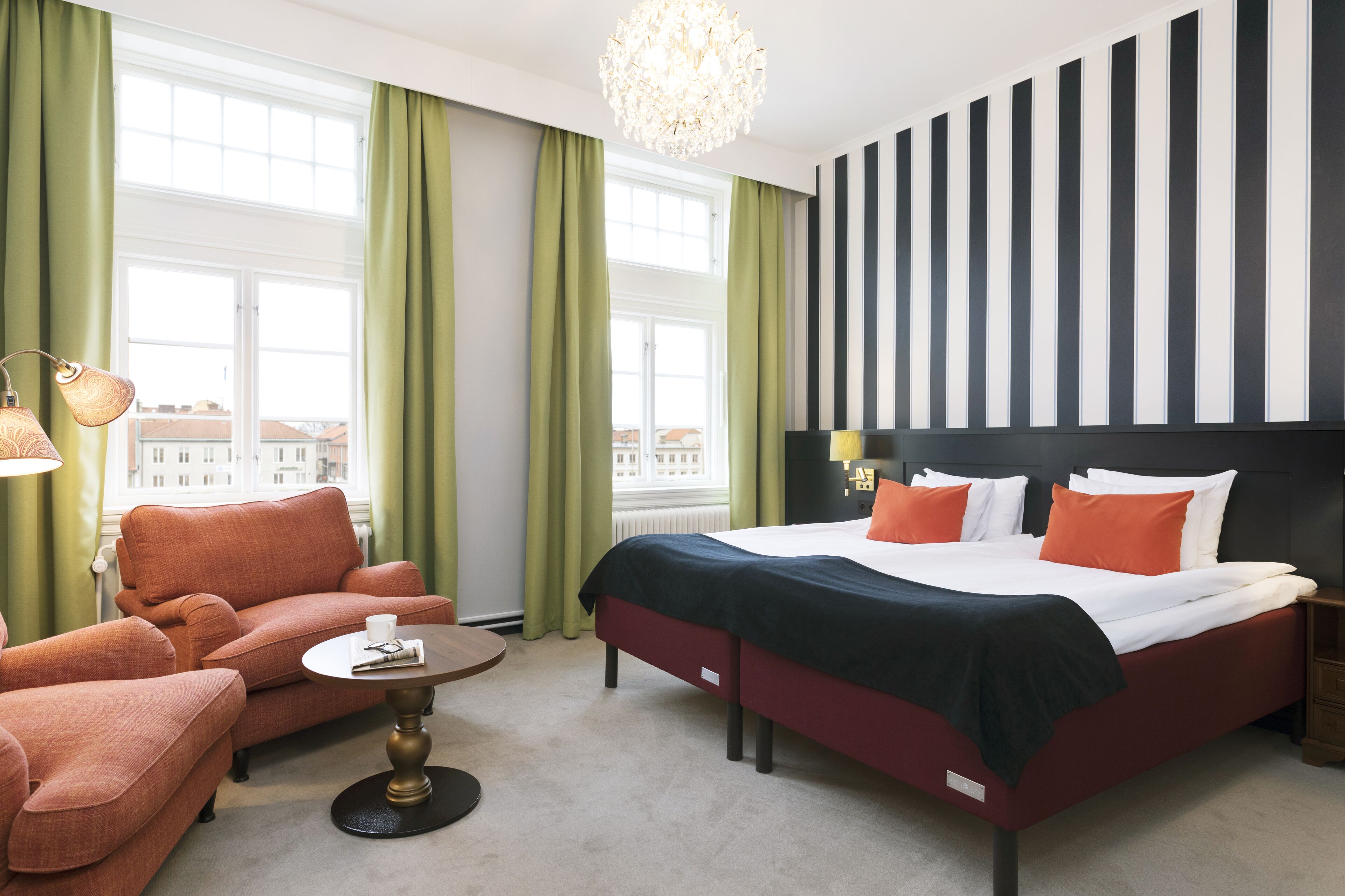 A hotel room with a large bed, a sofa, a small table, and a view of buildings through the window.