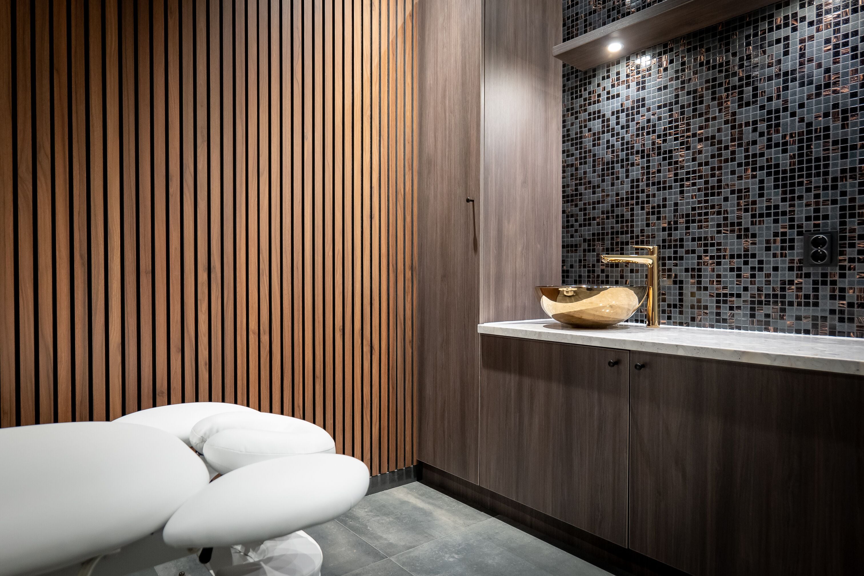 Un baño moderno con pared de listones de madera, salpicadero de mosaicos y un lavamanos blanco con grifo dorado.