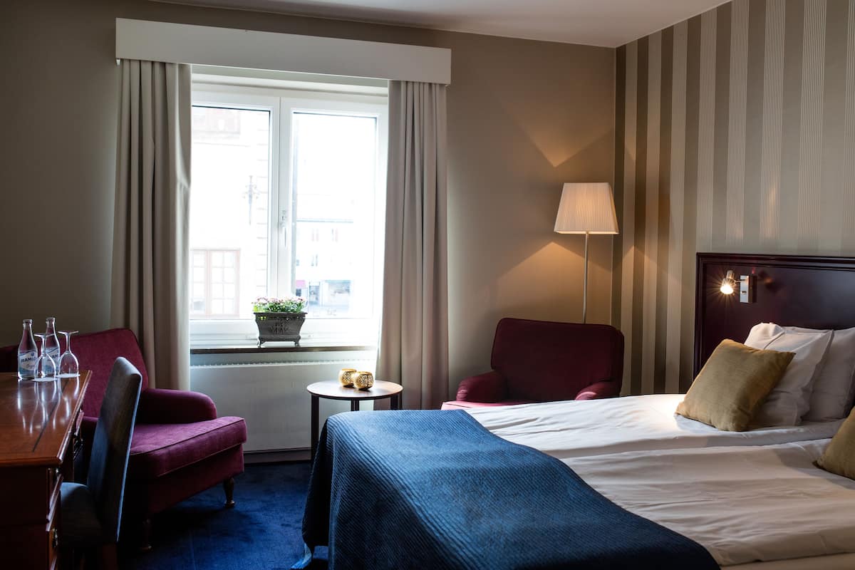 A hotel room with a bed, a desk with chairs, a window with curtains, and a lamp.