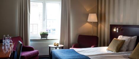 Una habitación de hotel con una cama, un escritorio con sillas, una ventana con cortinas y una lámpara.