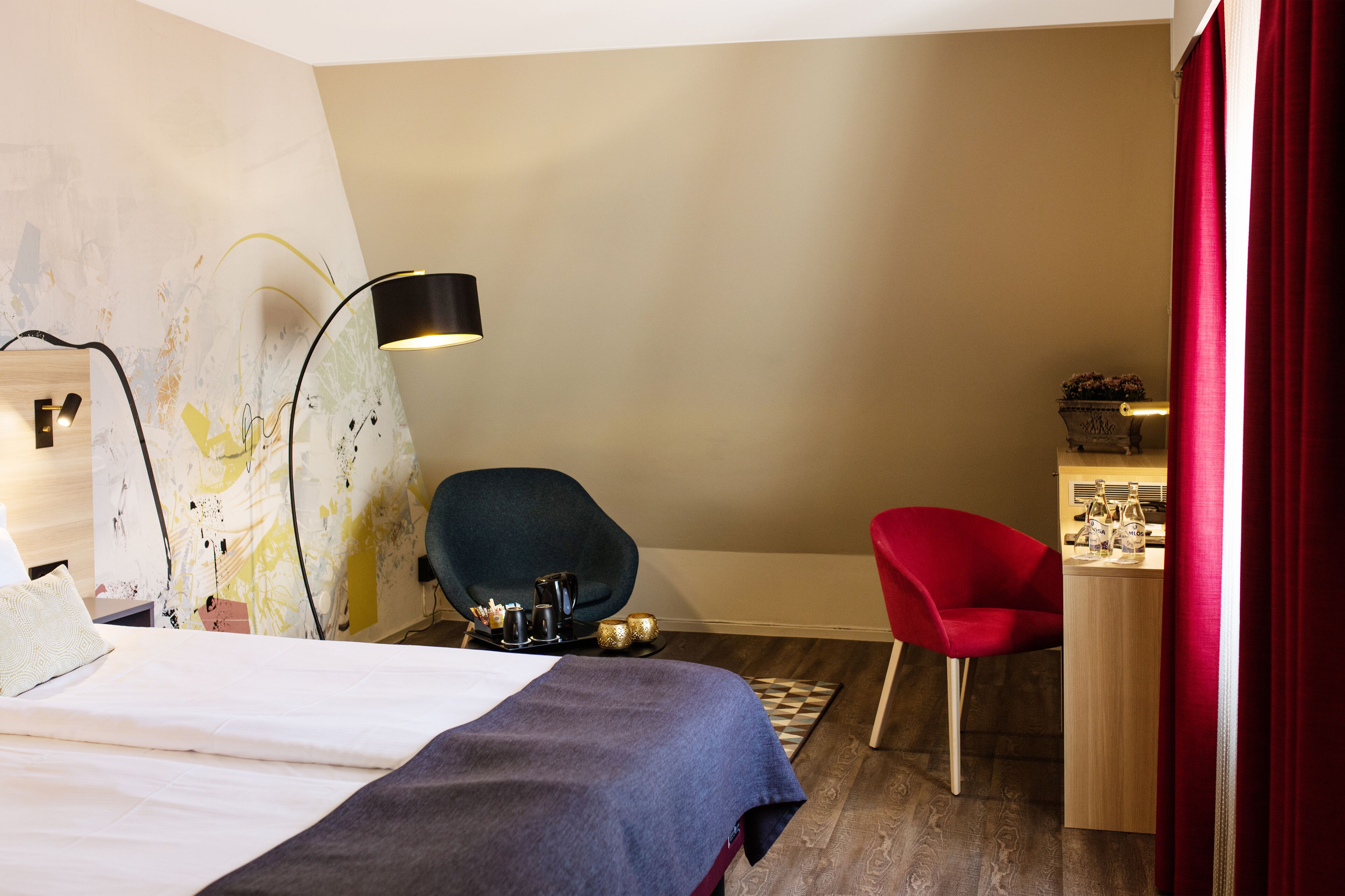 A hotel room with a bed, a desk with a chair, a lamp, and a small table with bottles.