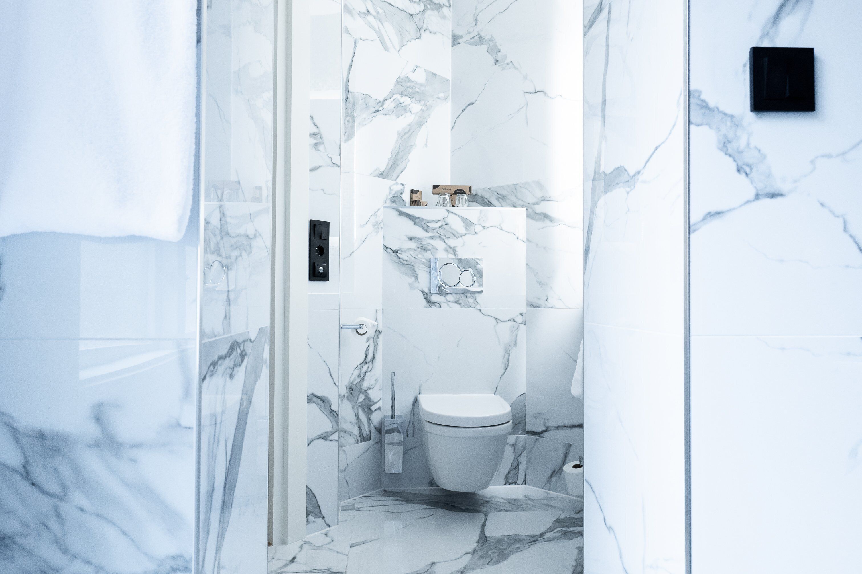 Une salle de bain avec un sol et des murs en marbre, des toilettes et une petite étagère contenant des articles de toilette.