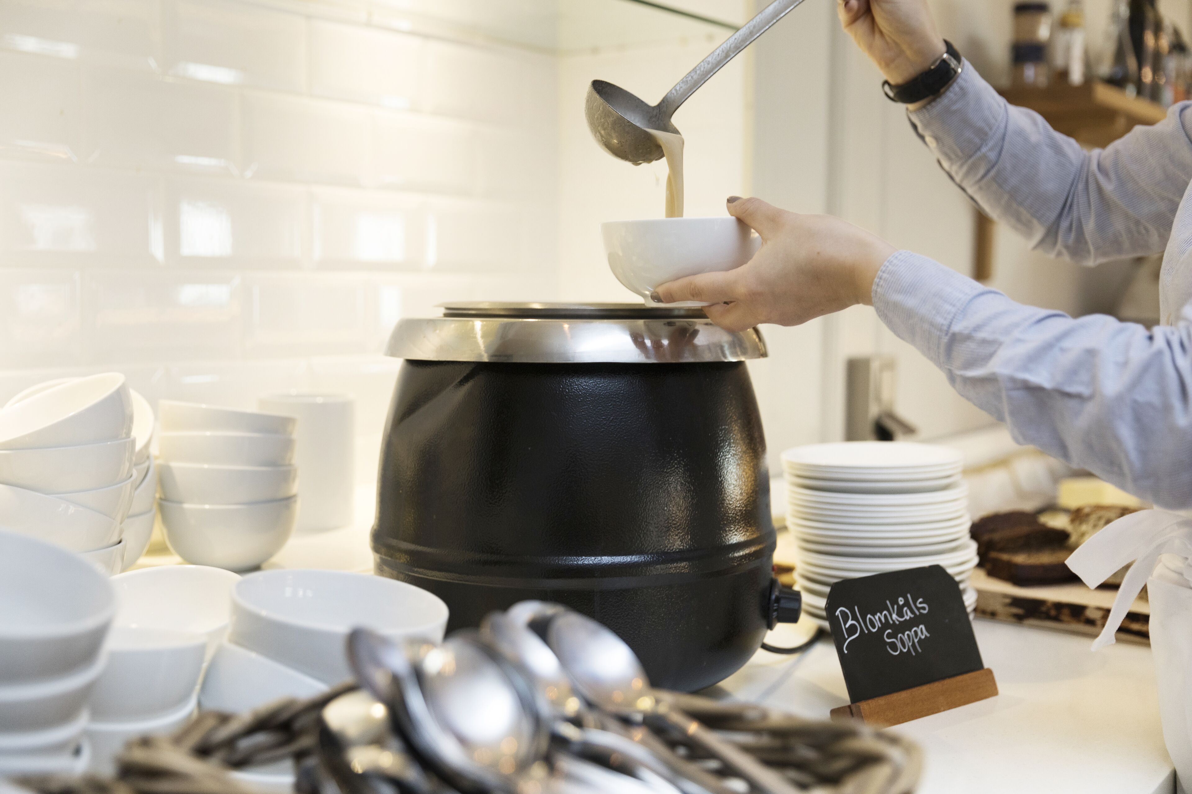 Una persona sirve sopa de una olla grande a un plato. Hay varios platos y cucharas sobre la mesa, y un letrero que dice “Blomkål's Suppa”.