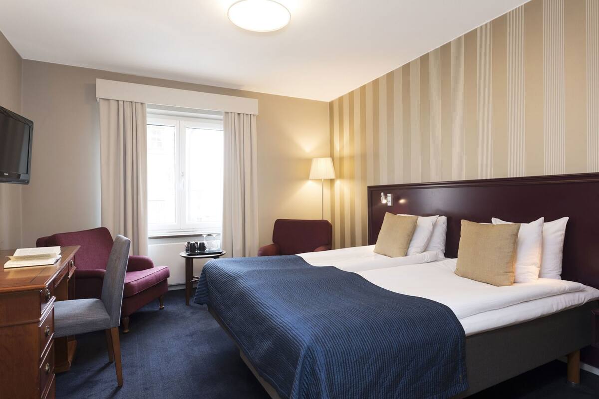 A hotel room with a bed, a desk, a chair, a lamp, and a TV.