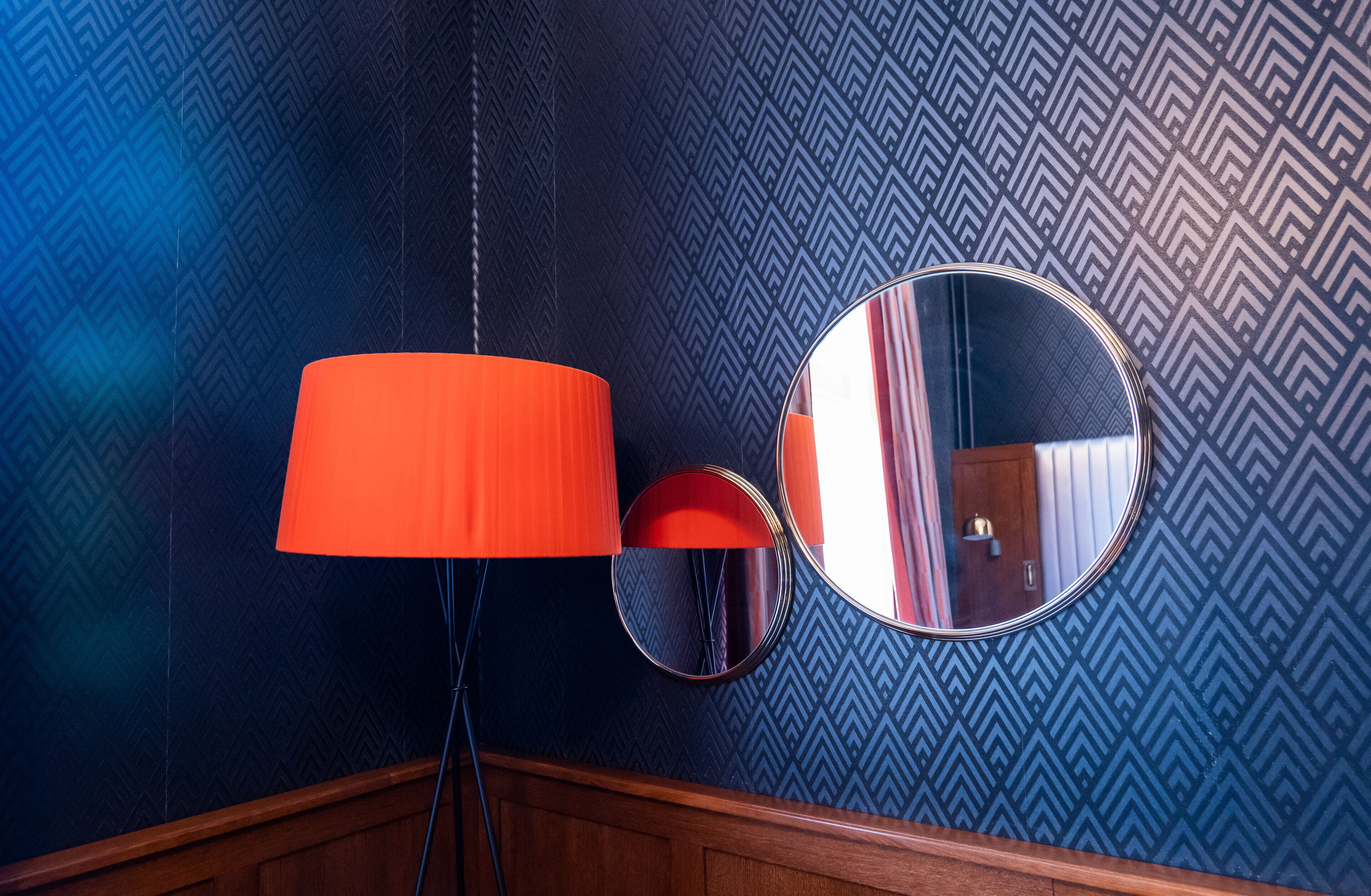 A room with a patterned wallpaper, a round mirror, and a floor lamp with an orange shade.