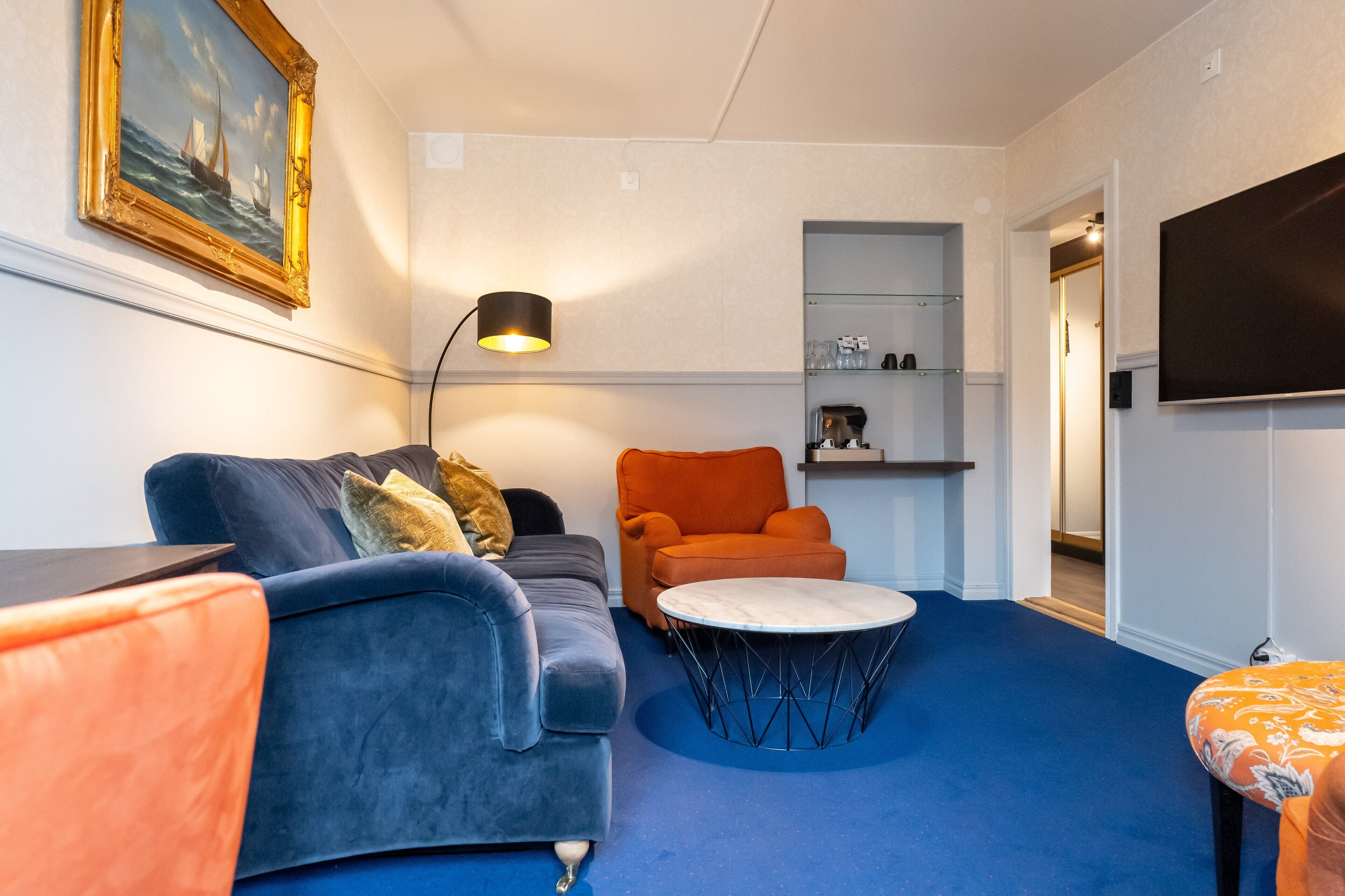 A modern living room with a blue carpet, a blue sofa, an orange armchair, a round coffee table, and a painting on the wall.
