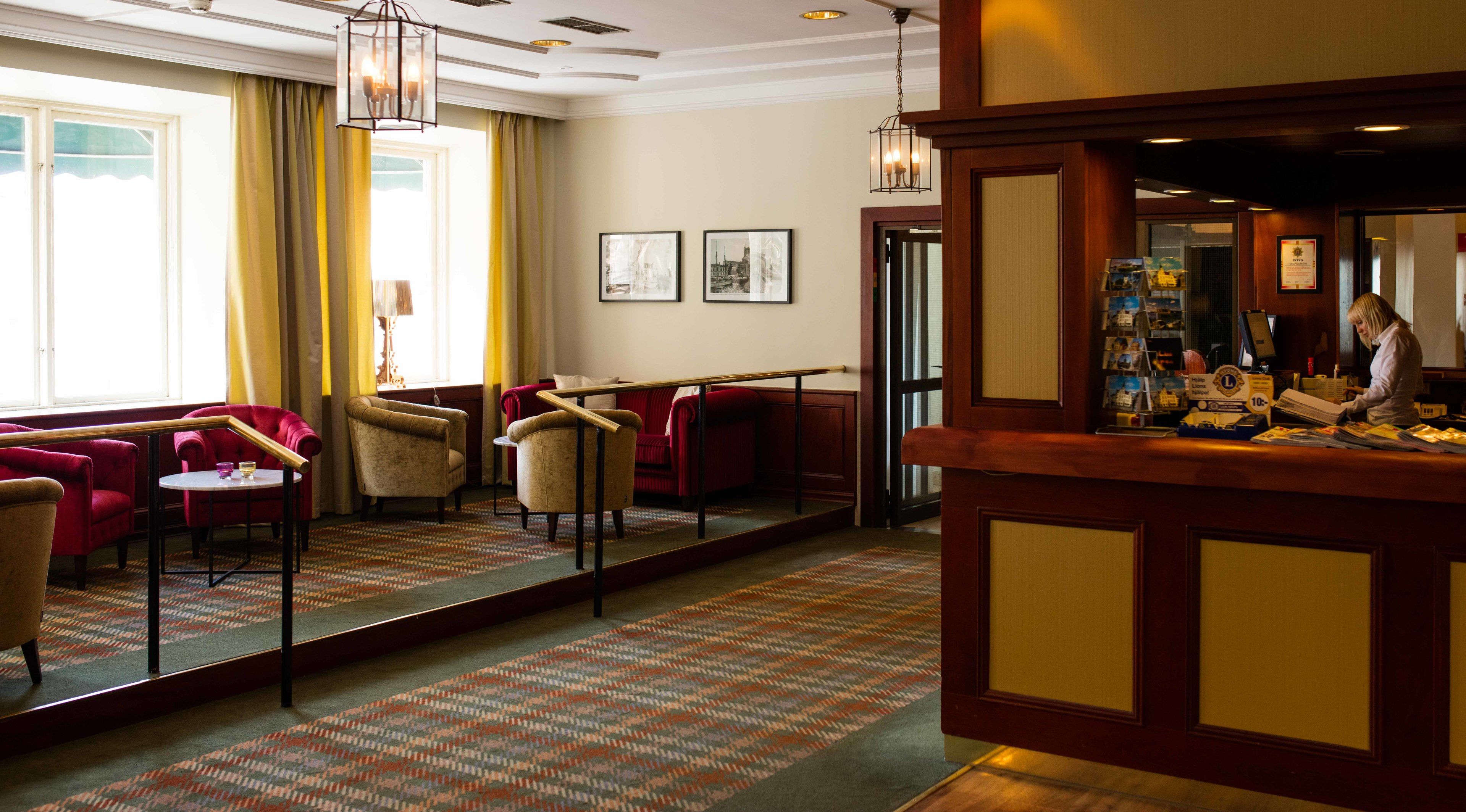 A hotel lobby with a reception desk, red and beige armchairs, a patterned carpet, and framed artwork on the wall.