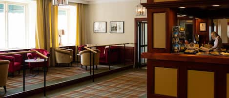 Um hall de hotel com uma recepção, poltronas vermelhas e bege, um tapete estampado e obras de arte emolduradas na parede.