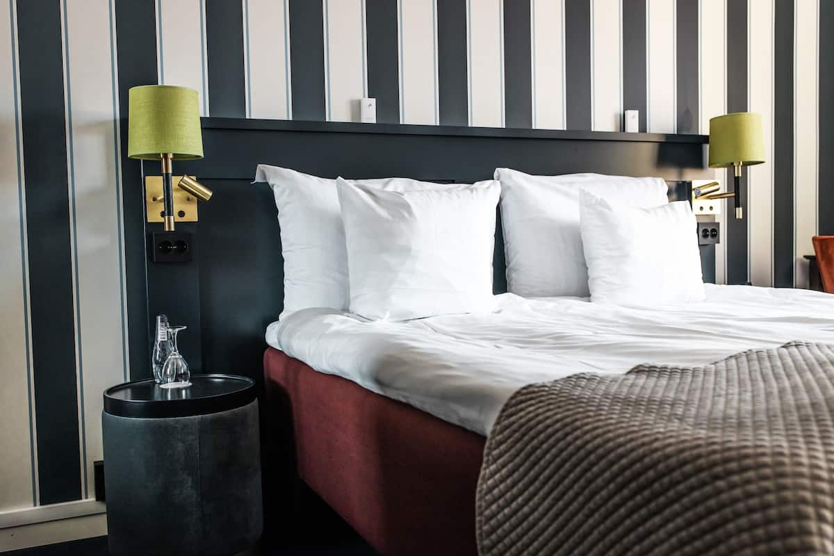 A hotel room with a bed, two bedside lamps, a bedside table with a glass pitcher, and a red upholstered bench.