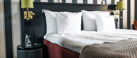 A hotel room with a bed, two bedside lamps, a bedside table with a glass pitcher, and a red upholstered bench.
