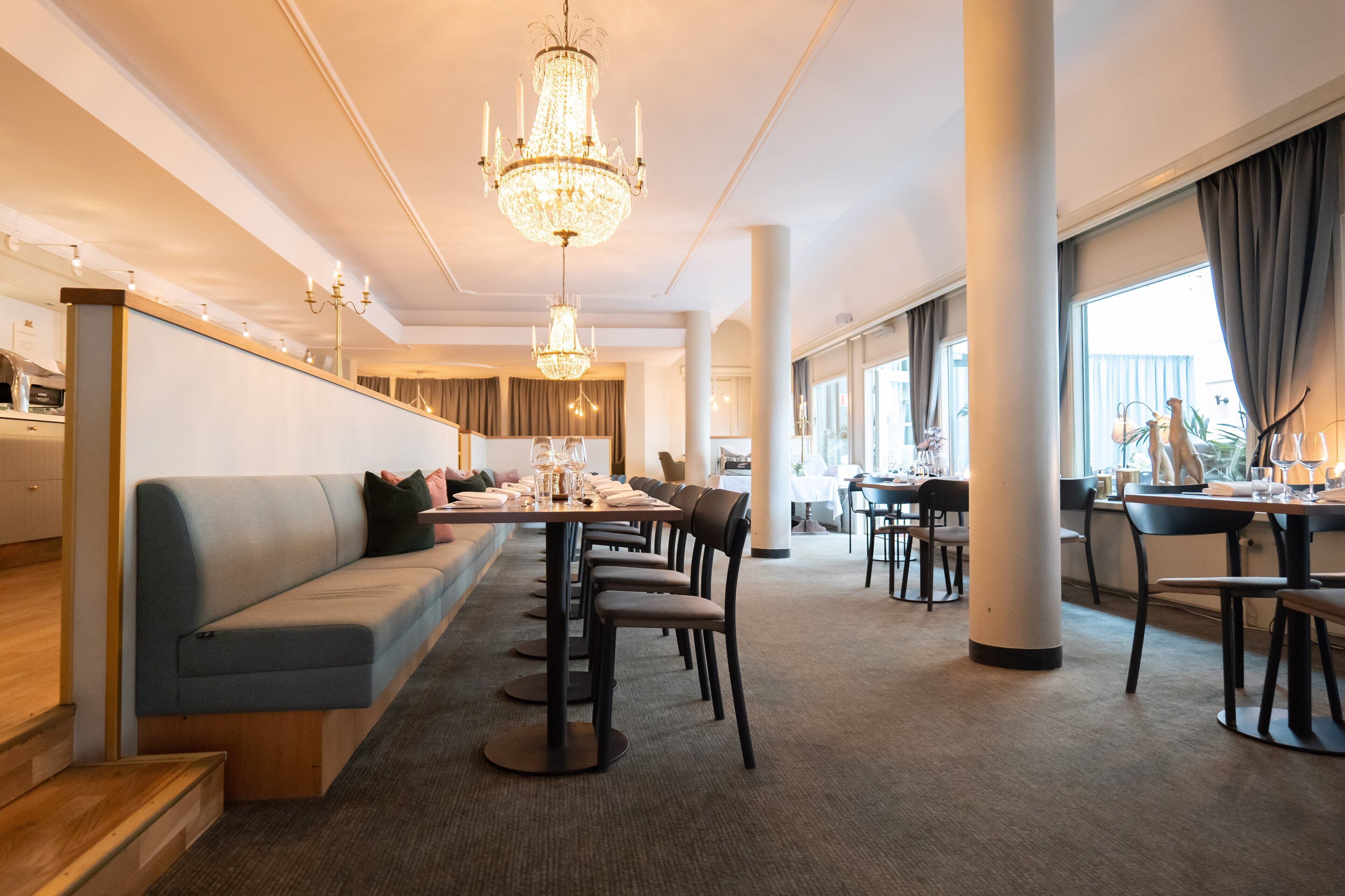 A modern hotel dining area with a long communal table, several chairs, a blue sofa, and a chandelier.