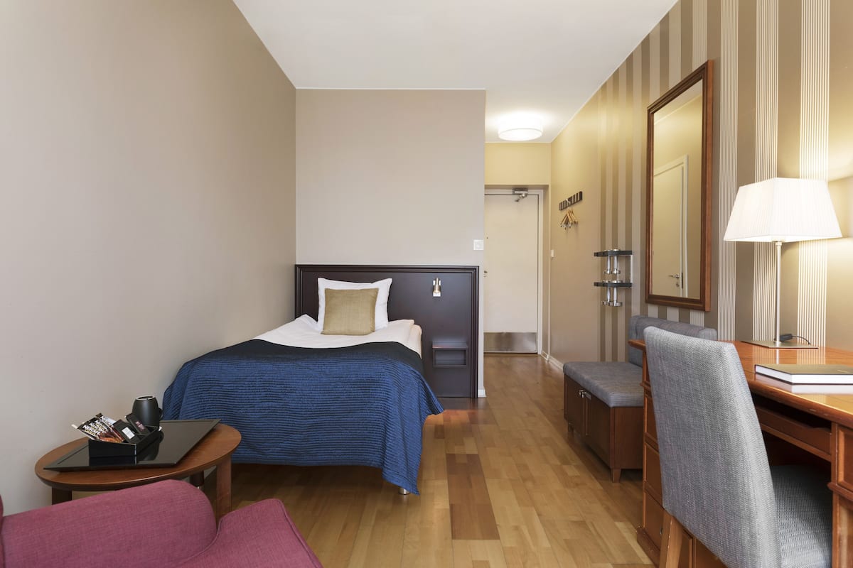 A hotel room with a bed, a desk, a chair, a small table, and a lamp.