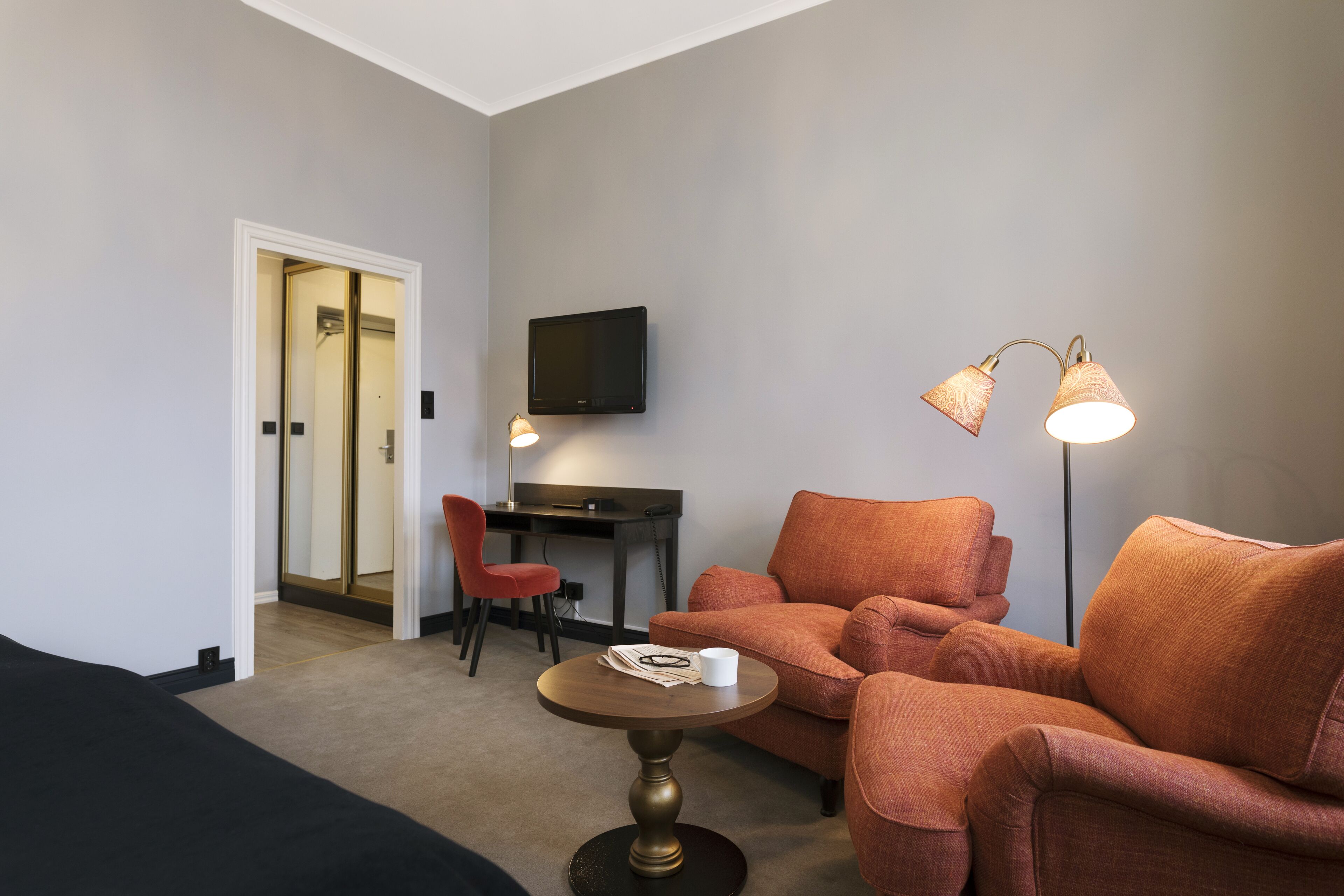 A hotel room with a sofa, a small round table, a desk with a chair, a TV mounted on the wall, and a floor lamp.