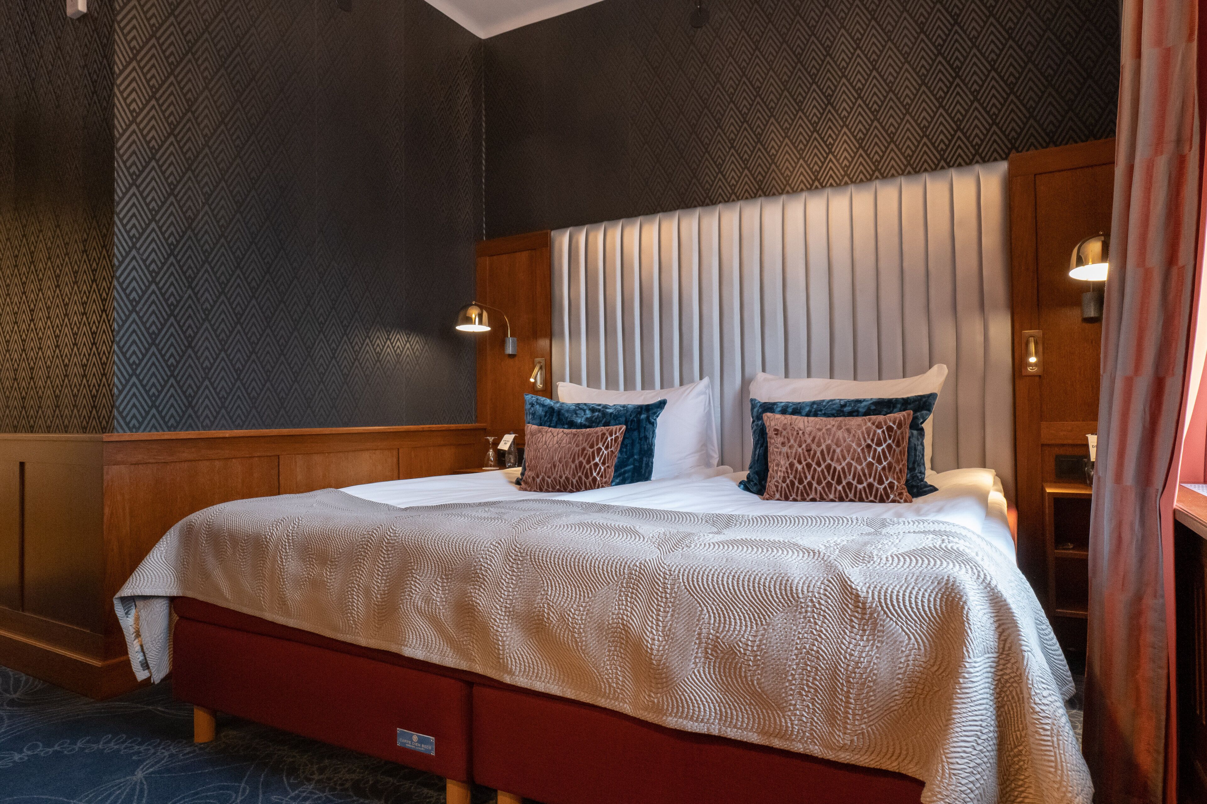 A neatly made bed with a patterned bedspread and two decorative pillows, a wooden headboard, and a wall with a geometric pattern.
