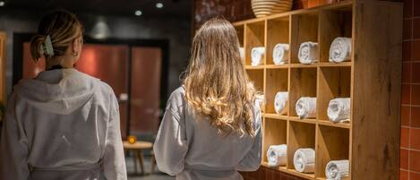 Duas pessoas em um quarto com uma prateleira de madeira com toalhas enroladas.