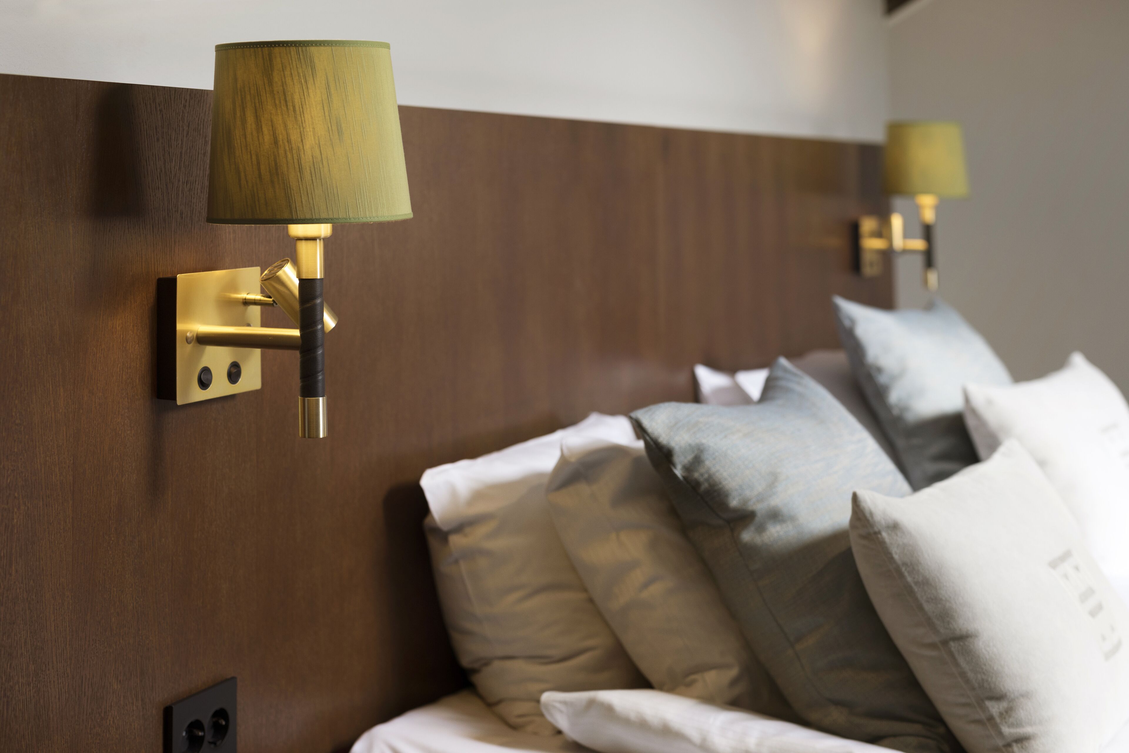 A hotel room with a wooden headboard, a bed with multiple pillows, and wall-mounted lamps.