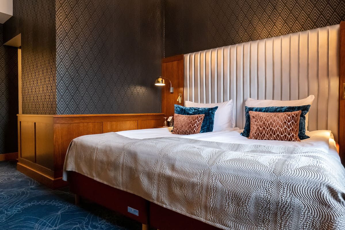 A hotel room with a large bed, a wooden headboard, a patterned wall, and a nightstand with a lamp.