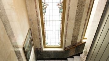 Uma escadaria de mármore com corrimão, que leva a um quarto com uma grande janela arqueada e um quadro na parede.