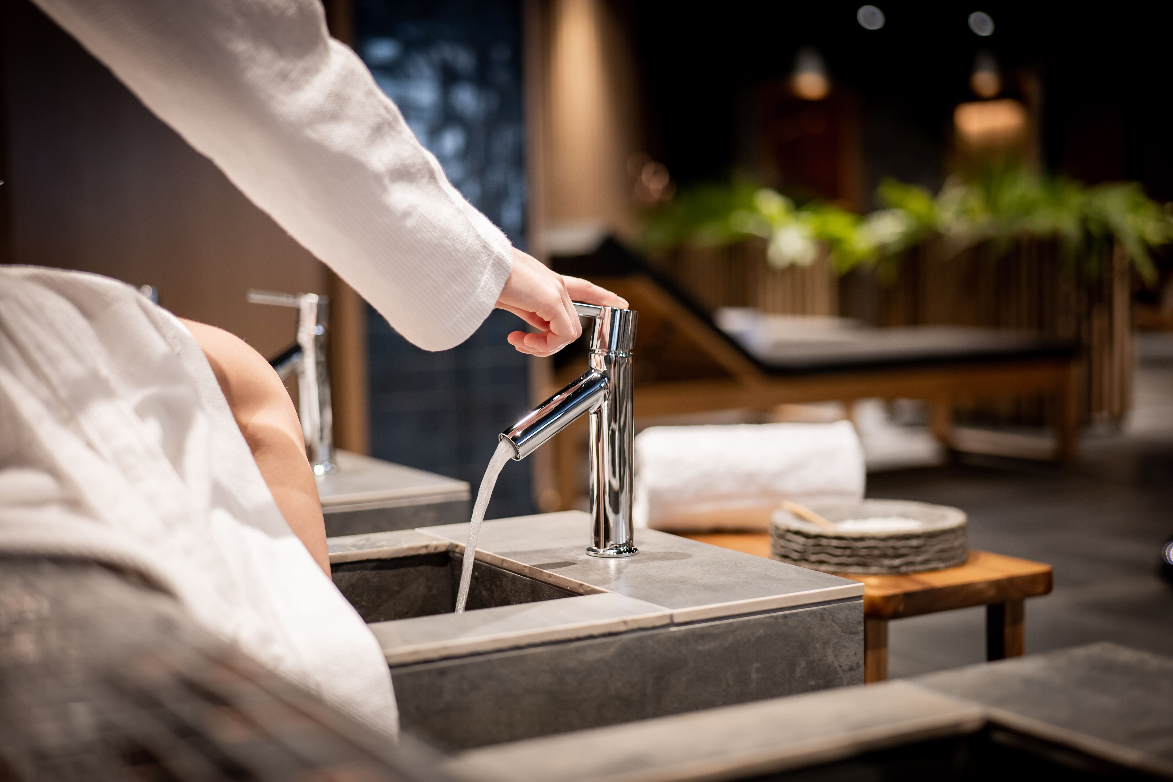 Una persona ajustando un moderno grifo en un baño con un lavabo de piedra y una mesa de madera con una toalla y una bandeja redonda.