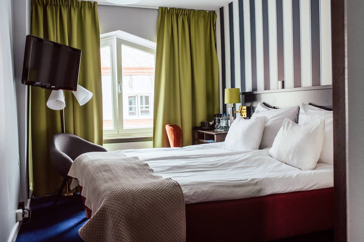 A hotel room with a bed, a chair, a desk with a lamp, and a television mounted on the wall.