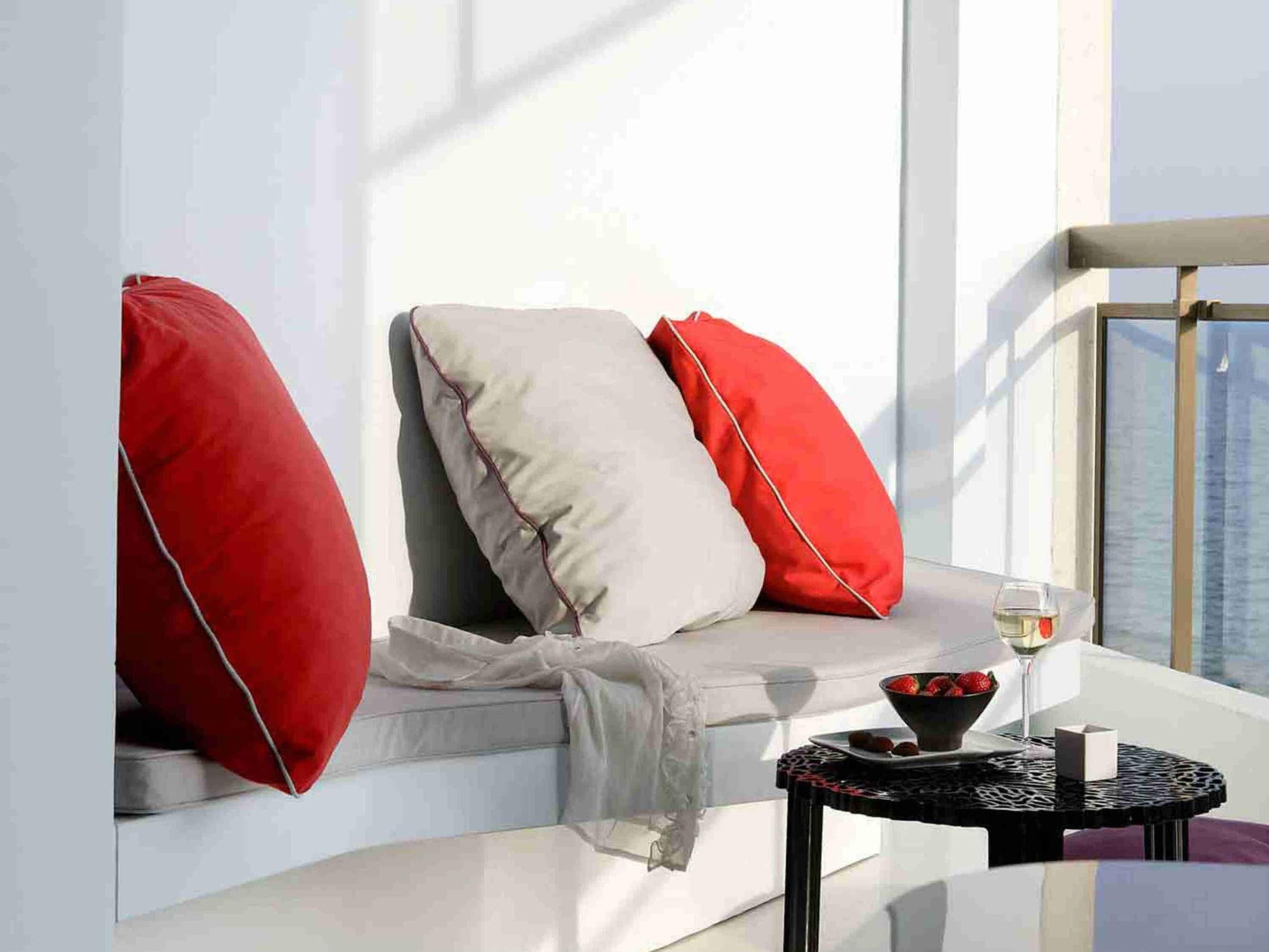 A modern balcony with a cushioned seating area, a small round table with a bowl of fruit and a glass of wine, and a view of the city skyline.