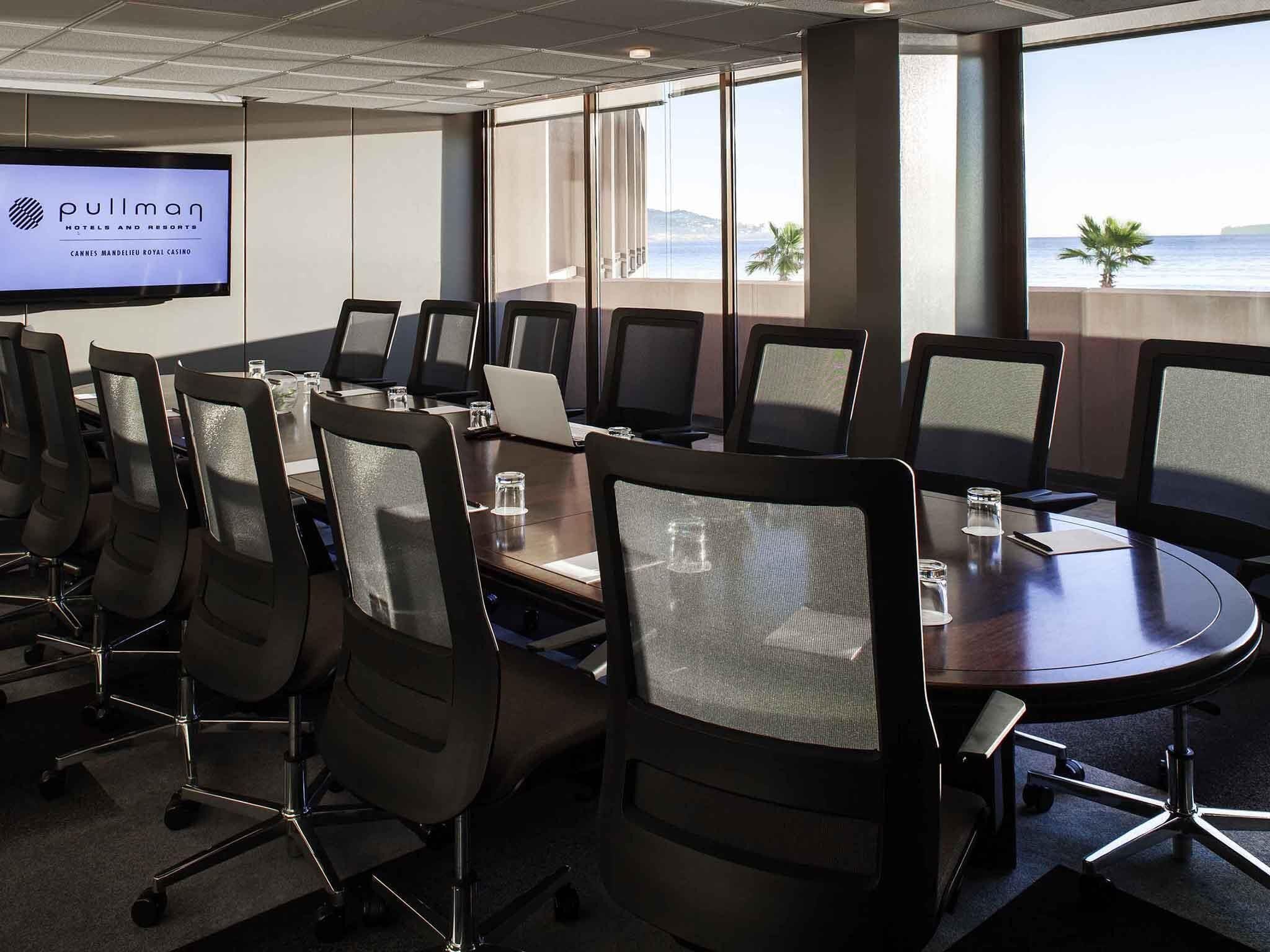 A conference room with a large screen displaying 'Pullman Hotels and Resorts' and 'Cannes Mandelieu Royal Casino.' The room has a modern design with glass partitions, a round table, and ergonomic chairs.