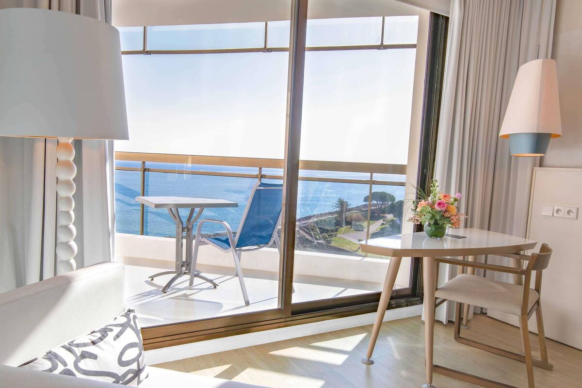 Un balcone con tavolo e sedie, vista sul mare e una finestra con tende.