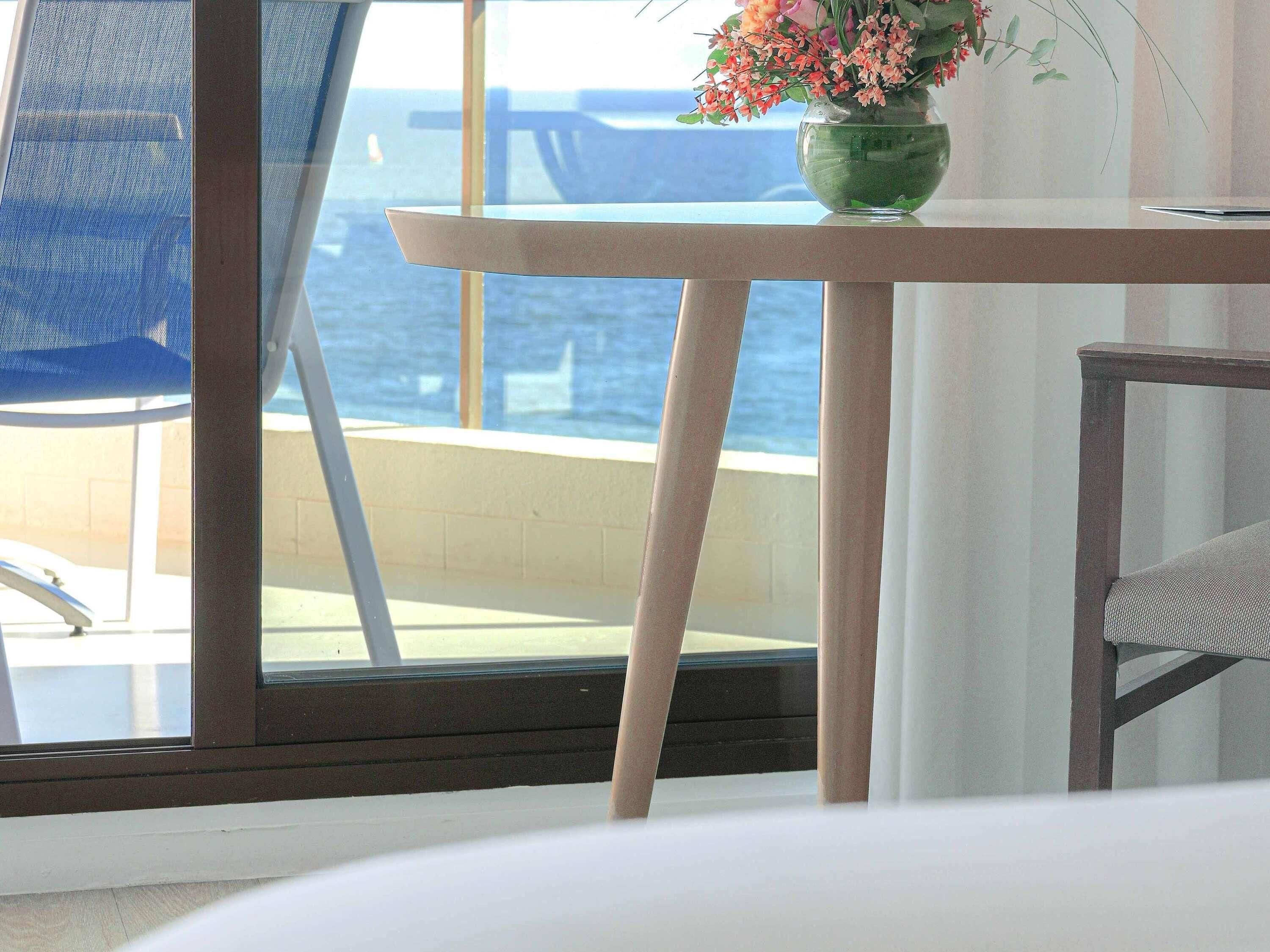 A room with a view of the sea, a table with a vase of flowers, and a chair.