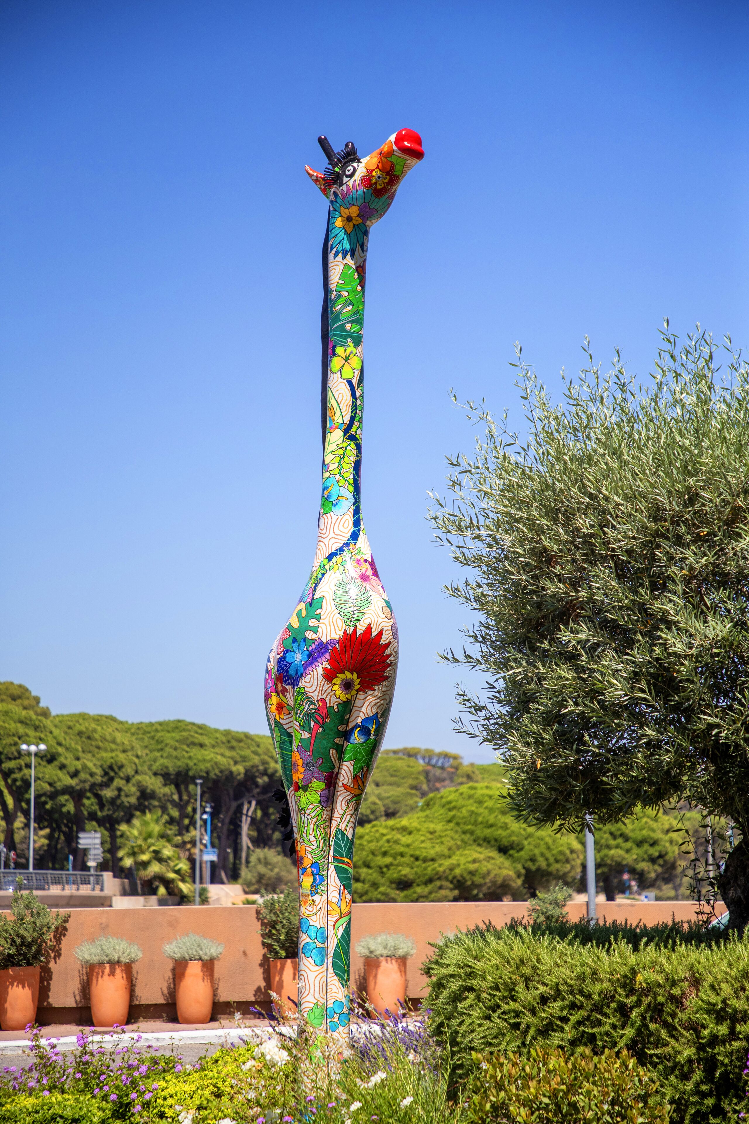 A tall, colorful sculpture of a giraffe in an outdoor setting with potted plants and trees.
