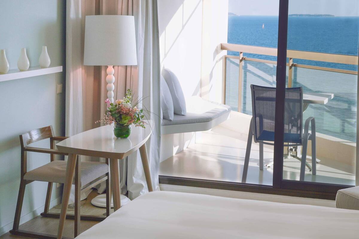 Camera d'hotel con balcone, tavolo con sedie, un letto e vista sul mare.