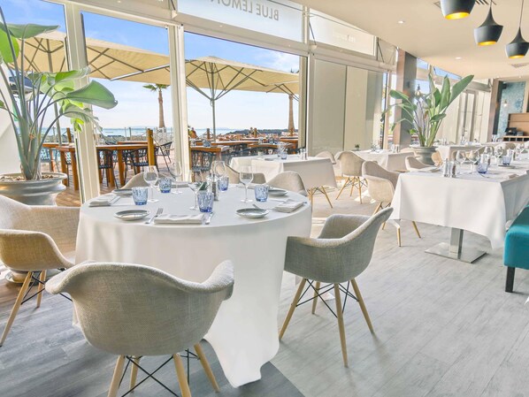 Un restaurante con mesas redondas dispuestas para comer, amplias ventanas con vista a la playa y plantas de interior.