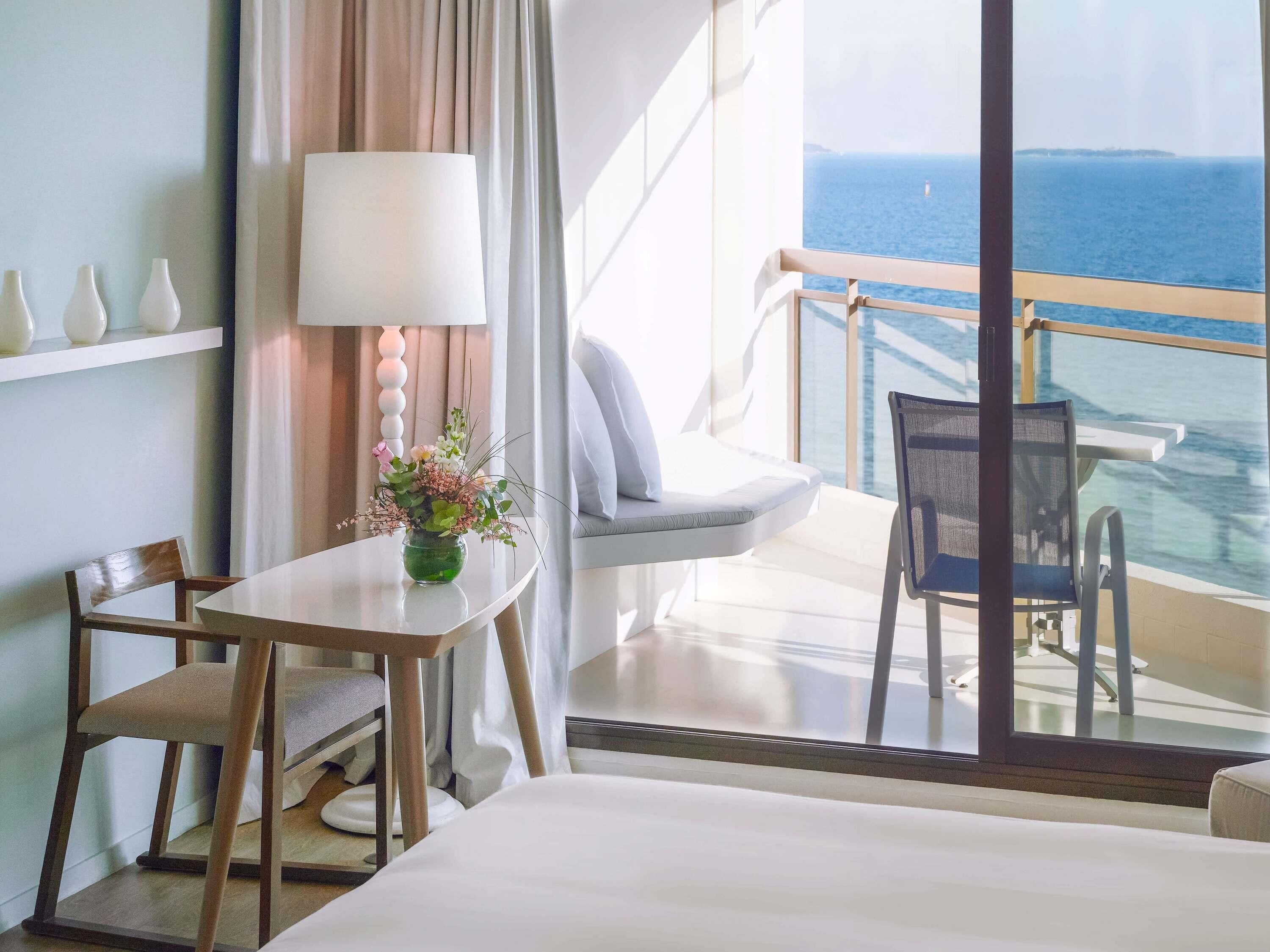 Habitación de hotel con cama, una mesa con un florero, una silla, balcón con vista al mar y una lámpara.