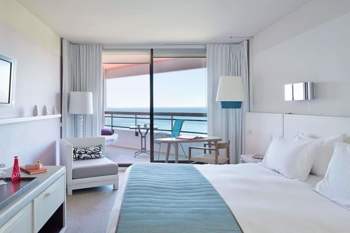 A hotel room with a bed, a desk, a chair, a small table, and a balcony with a view of the ocean.