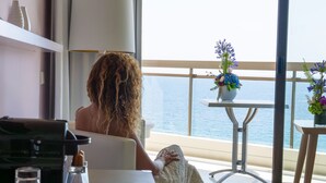 A room with a large window overlooking the sea, a small table with a vase of flowers, and a sofa with a cushion. - Pullman Cannes Mandelieu (Mandelieu-La-Napoule)