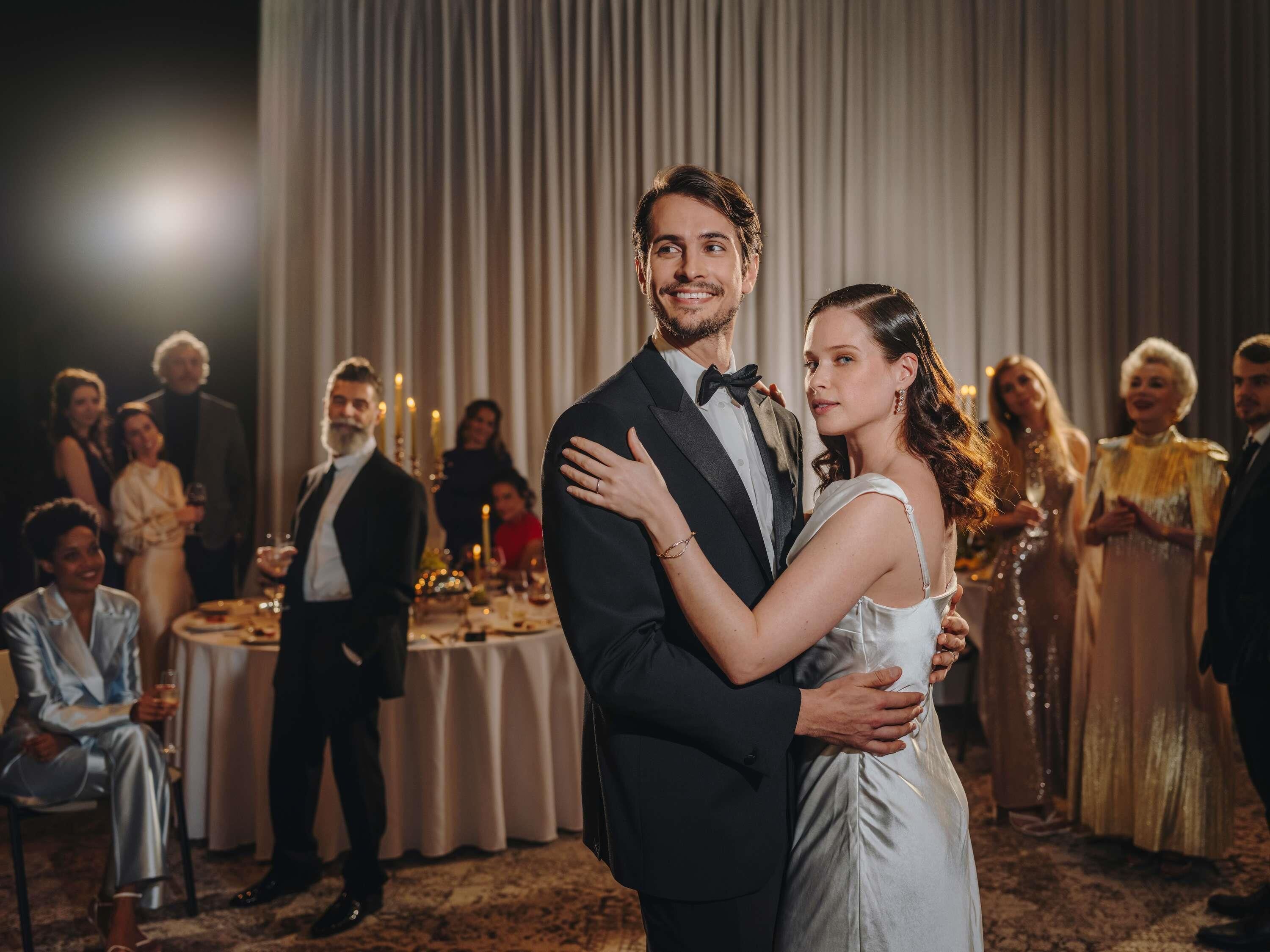A couple embracing in a formal event setting with a table and guests in the background.