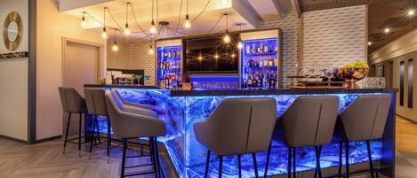 Una zona de bar con mostrador de mármol, taburetes, lámparas colgantes y estanterías llenas de botellas.